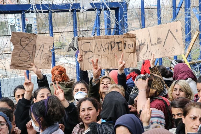 Sınır kapısında mülteci kadınlar ellerinde Türkçe, Yunanca ve Arapça 8 Mart yazılı dövüzler taşıyor.