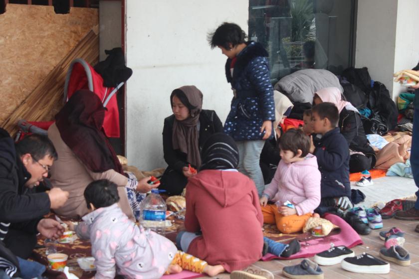 Yerde, betonda incecik battaniyenin üstünde yemek yiyen mülteci aile.