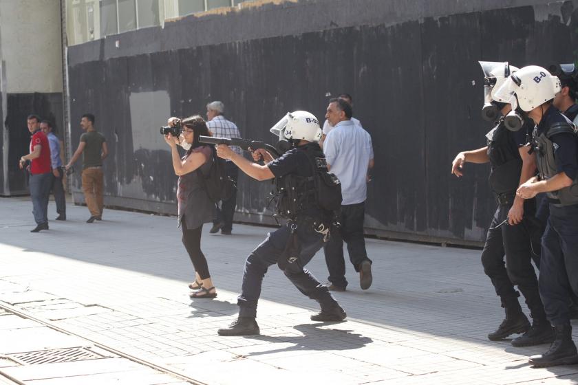 Yan yana duran polis ve gazeteci. Polis tetiğe, gazeteci deklanşöre basmak üzere. 