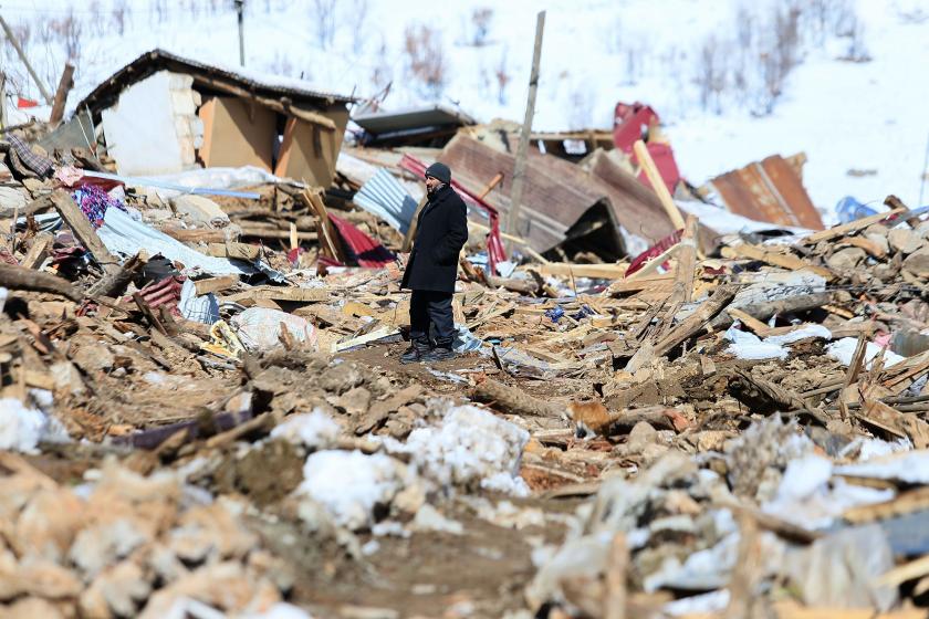 Depremin ardından enkazın üzerinde duran, siyah paltolu, bereli, orta yaşlı bir adamın fotoğrafı