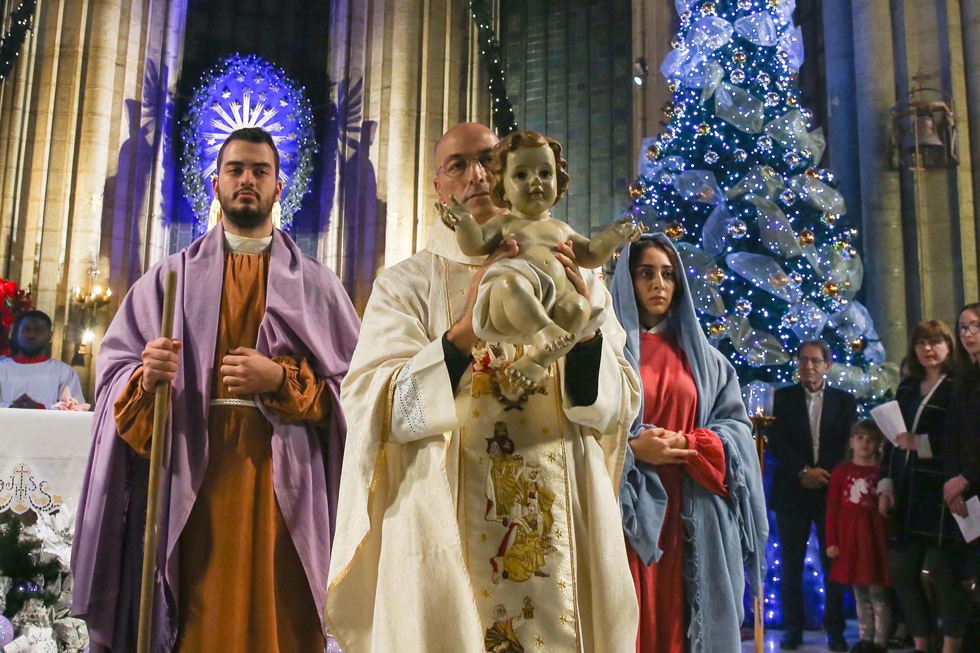 Saint Antuan Kilisesi'nde rahip elinde İsa'yı sembolize eden bebek heykeli tutuyor. Yanıbaşında genç bir erkek ve genç bir kadın var.