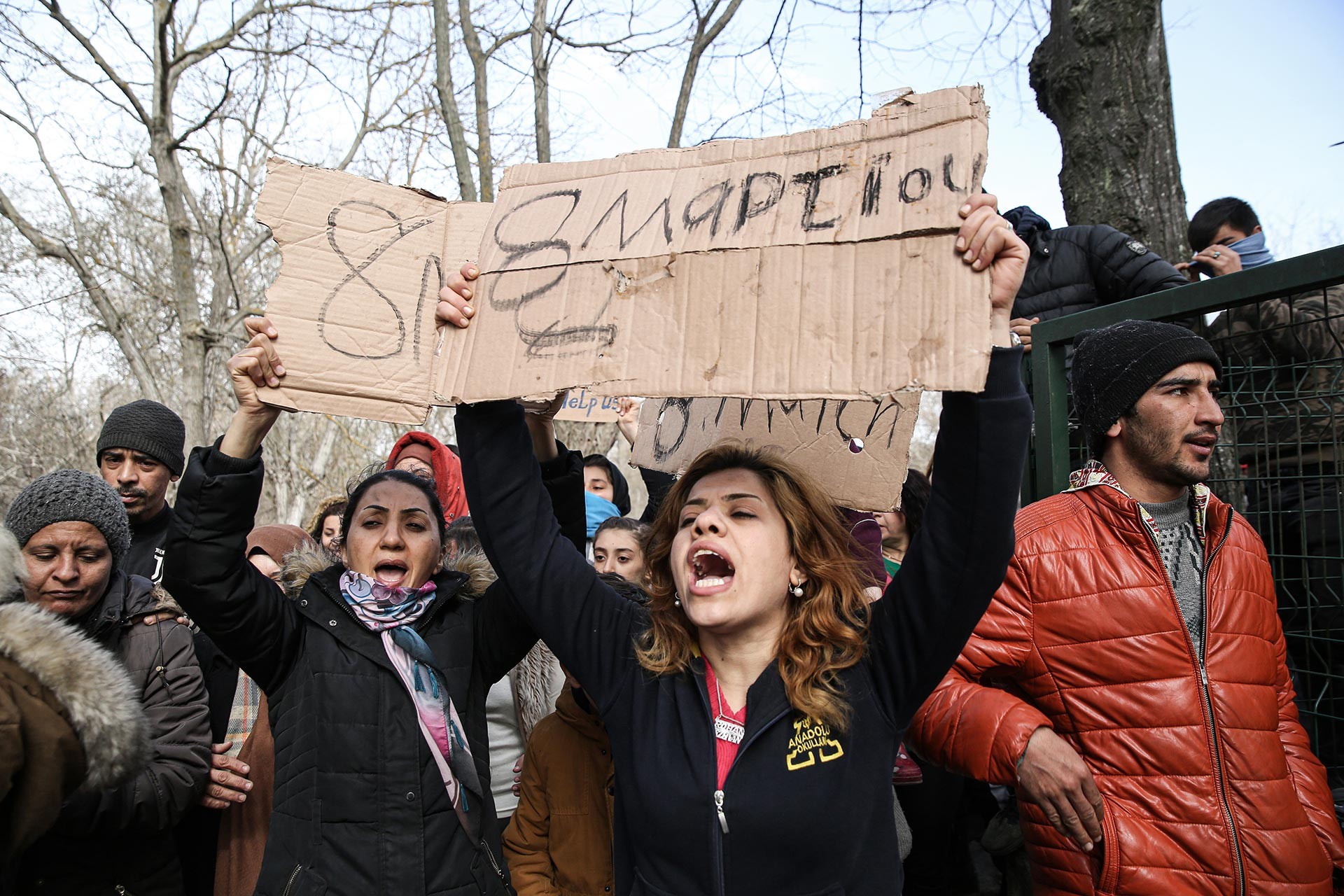 Sınır kapısında mülteci kadınlar ellerinde Türkçe, Yunanca ve Arapça 8 Mart yazılı dövüzler taşıyor.