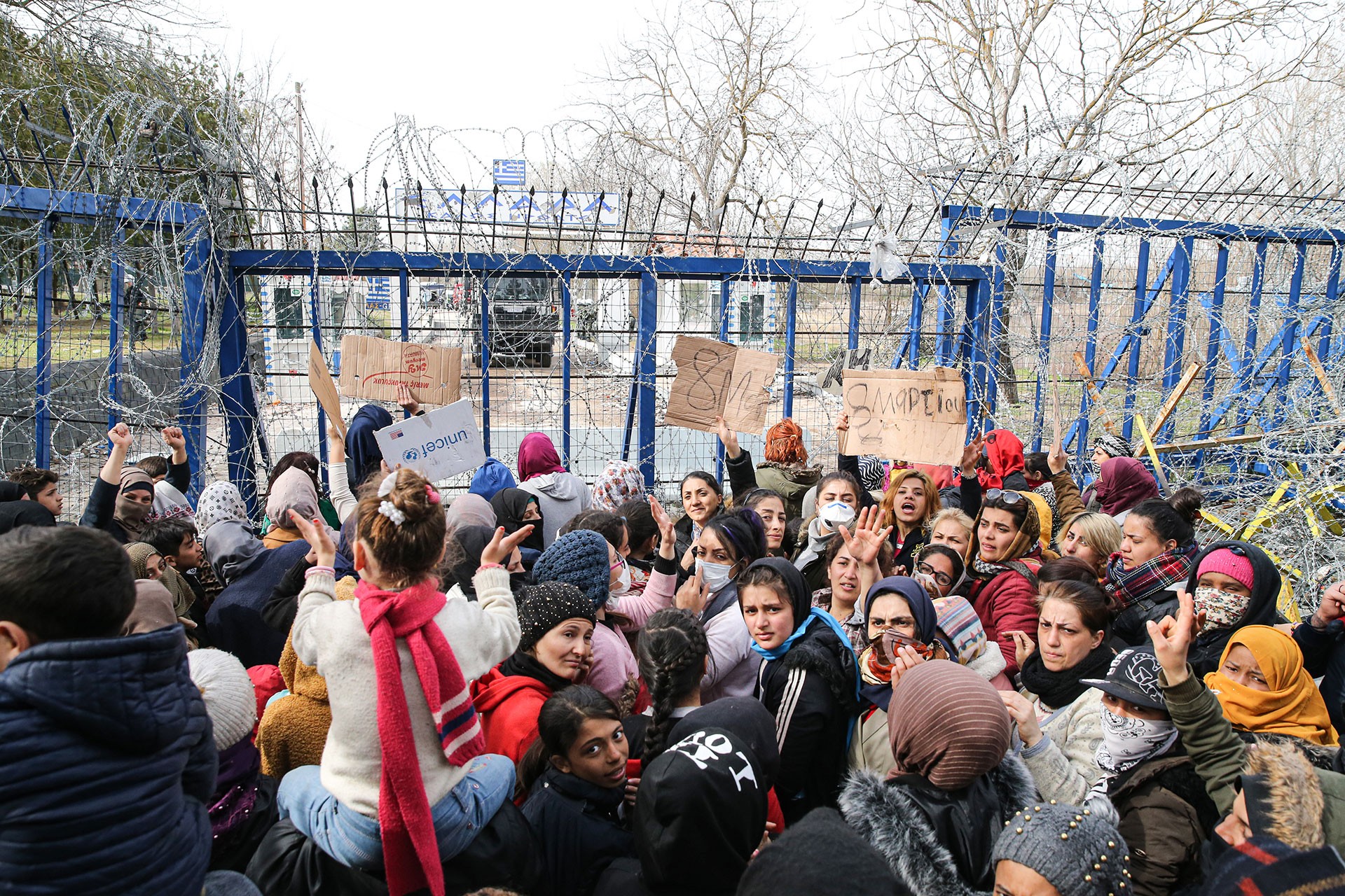 Sınır kapısında mülteci kadınlar ellerinde Türkçe, Yunanca ve Arapça 8 Mart yazılı dövüzler taşıyor.