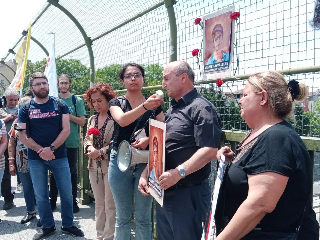 elnde karanfil, bayrak ve Mehmet Ayvalıtaş'ın fotoğrafları olan insanlar